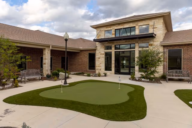 Outdoor courtyard area at Sedona Trace Health & Wellness featuring a small artificial putting green with two golf flags, surrounded by concrete walkways, benches, plants, and a brick and stone building with large windows and a glass door under a cloudy sky.