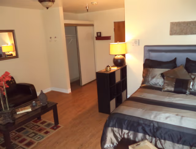 A cozy bedroom in an assisted living facility featuring a bed with striped bedding and multiple pillows, a bedside table with a lamp, a small black shelving unit, a black armchair next to a coffee table with decorative items, a mirror on the wall, and a closet with sliding doors. The room has wooden flooring and neutral-colored walls.