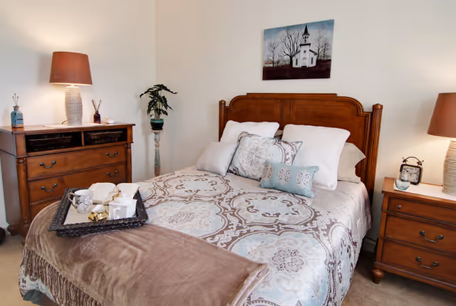 A cozy bedroom with a wooden headboard bed dressed in patterned bedding and multiple pillows. There is a brown throw blanket at the foot of the bed with a tray holding a teapot, cups, and a book. On either side of the bed are wooden nightstands with matching lamps. A small plant stands on a pedestal in the corner, and a painting of a white church hangs on the wall above the bed.