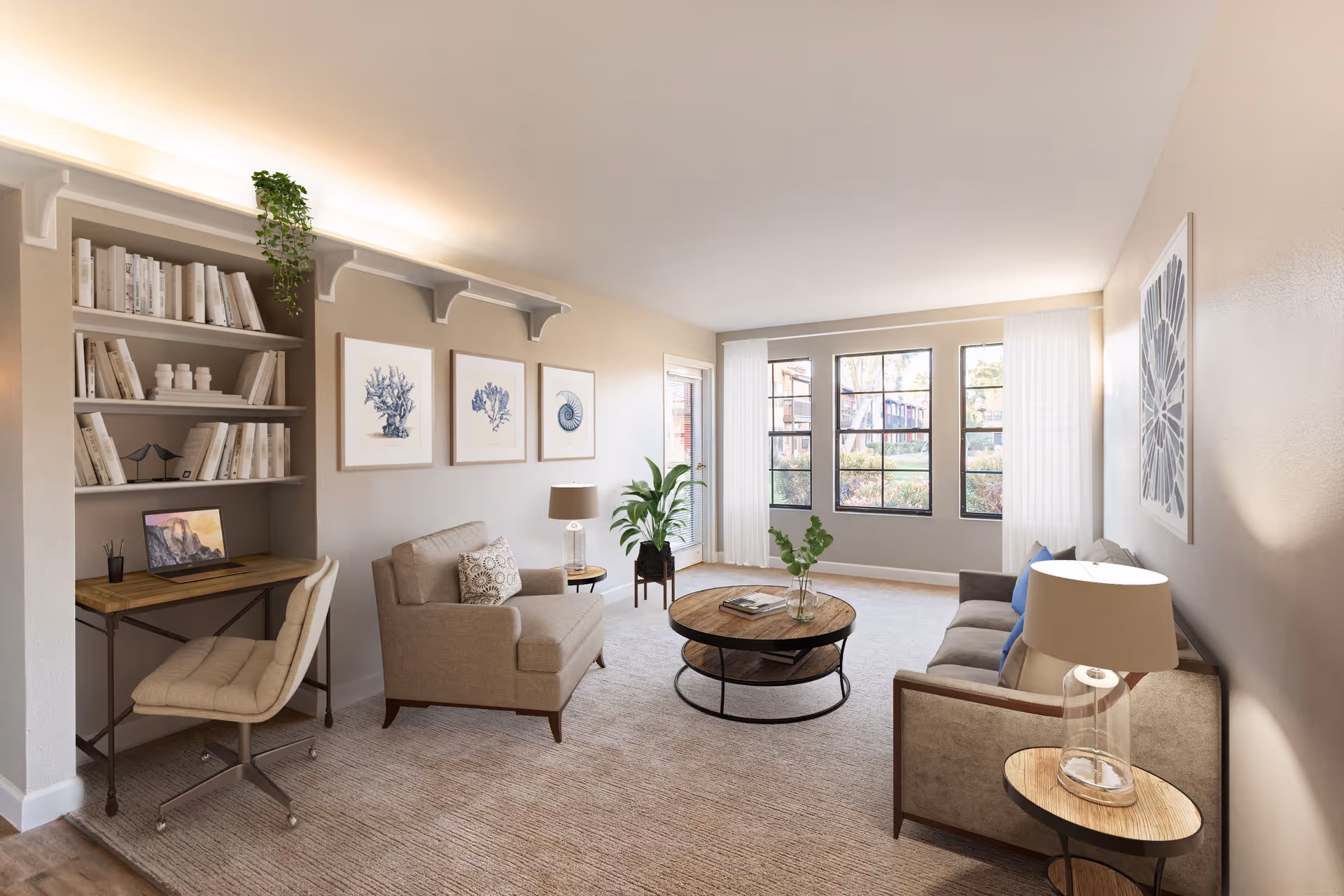 A bright and cozy living room in a senior living facility featuring a beige armchair and sofa with blue and patterned cushions, a round wooden coffee table with a glass vase and books, a small side table with a lamp, large windows with white curtains letting in natural light, and a built-in desk with a chair and laptop under shelves filled with books and decorative items. The walls are adorned with framed botanical artwork and a large floral print.