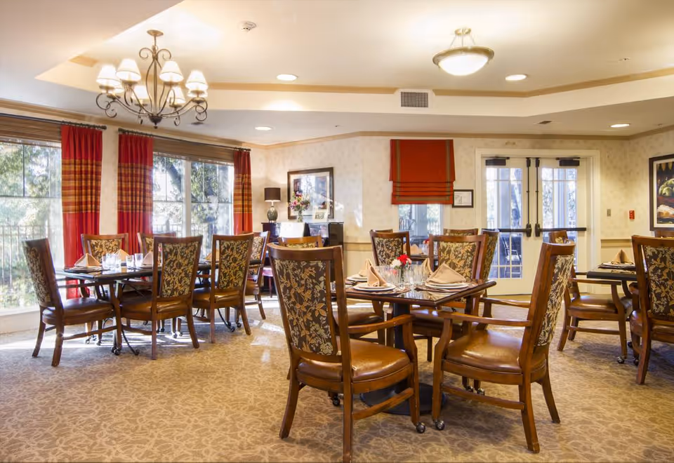 A bright dining room with multiple wooden tables and chairs arranged neatly. Each table is set with folded napkins, glasses, and small floral centerpieces. Large windows with red and plaid curtains allow natural light to fill the room. The walls are decorated with framed artwork and a sideboard with a lamp and flowers. The ceiling features a chandelier and recessed lighting.