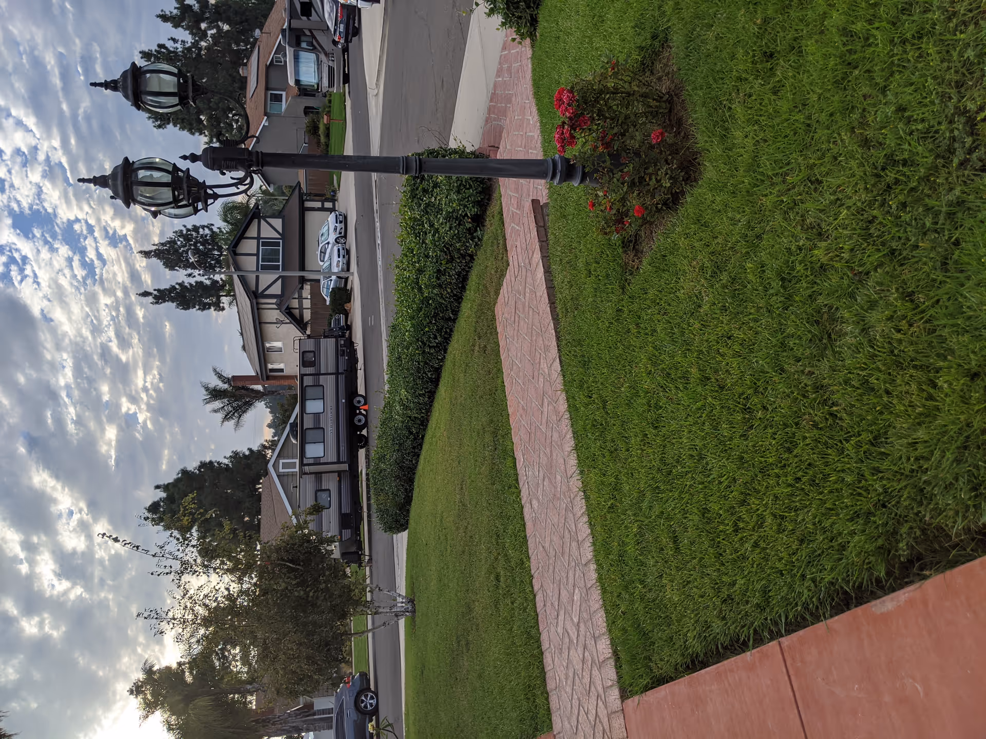 A suburban neighborhood street view with green lawns, a brick walkway, a black vintage-style street lamp with red flowers at its base, several houses, parked cars, and a trailer. The sky is partly cloudy with sunlight breaking through.