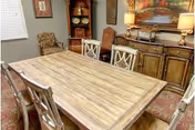 A dining room with a large wooden dining table surrounded by six chairs. The room features a sideboard with two lamps and decorative items, a large painting on the wall, and two upholstered armchairs in the corner. The floor is covered with a patterned rug, and there is a window with blinds on the left side.