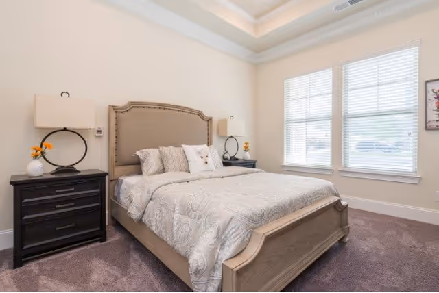 A bright and neatly arranged bedroom featuring a large bed with a beige upholstered headboard, patterned bedding, and multiple pillows. On each side of the bed are dark wooden nightstands with modern lamps and decorative items. The room has light-colored walls, a tray ceiling with recessed lighting, and two large windows with white blinds allowing natural light to enter.