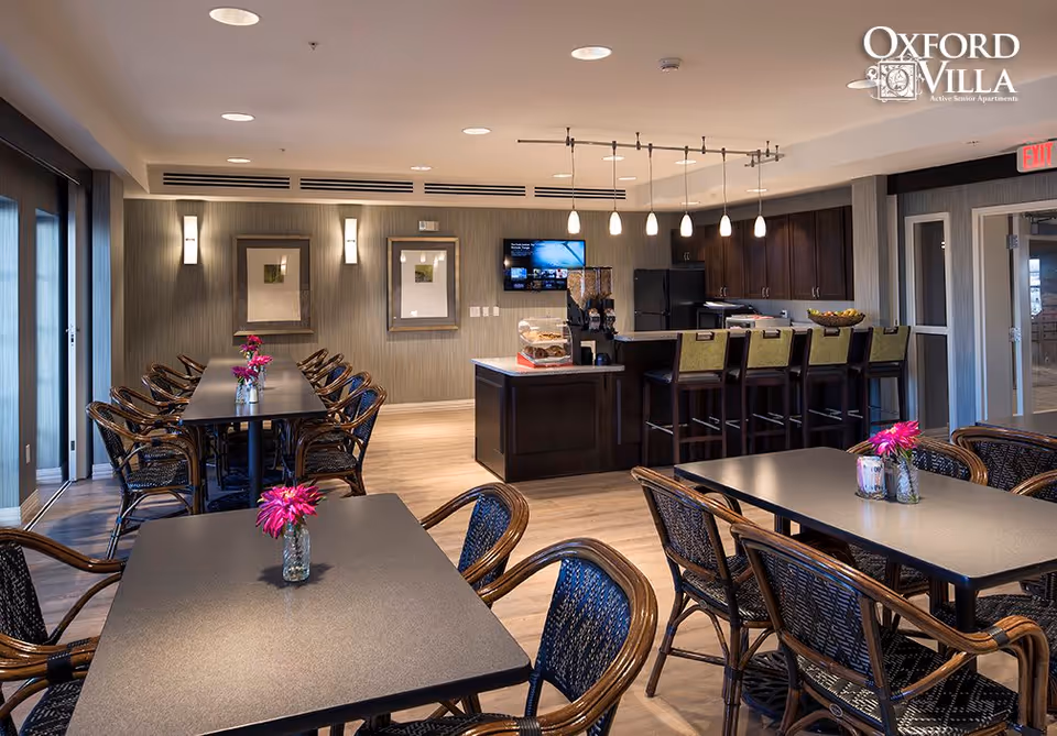 A modern dining area in Oxford Villa Active Senior Apartments featuring multiple tables with wicker chairs and small flower vases. In the background, there is a kitchen area with a breakfast bar, high chairs, pendant lighting, and a wall-mounted TV. The room has wooden flooring, framed artwork on the walls, and large windows letting in natural light.