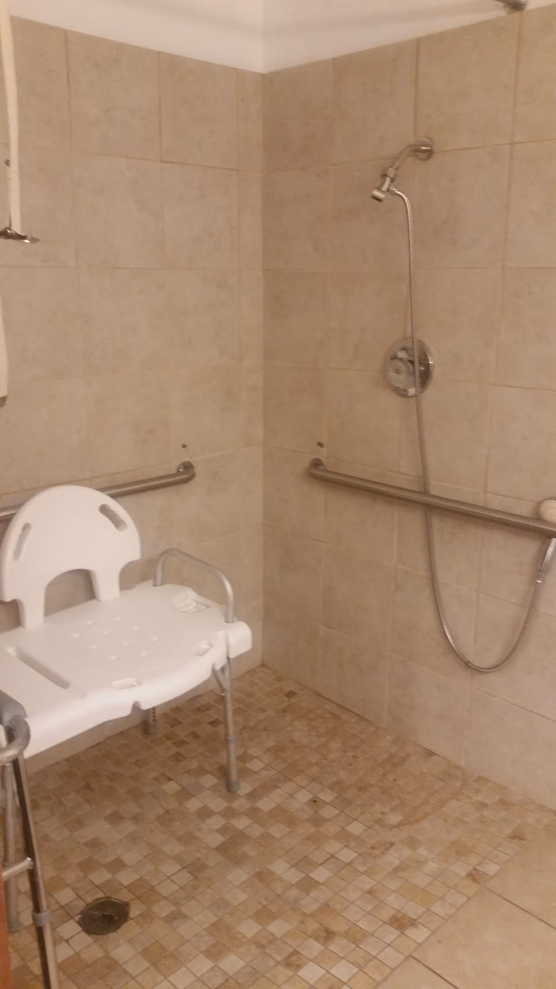 Accessible shower area with beige tiled walls and floor, equipped with a white plastic shower chair and metal grab bars on the walls. A handheld showerhead is mounted on the wall.