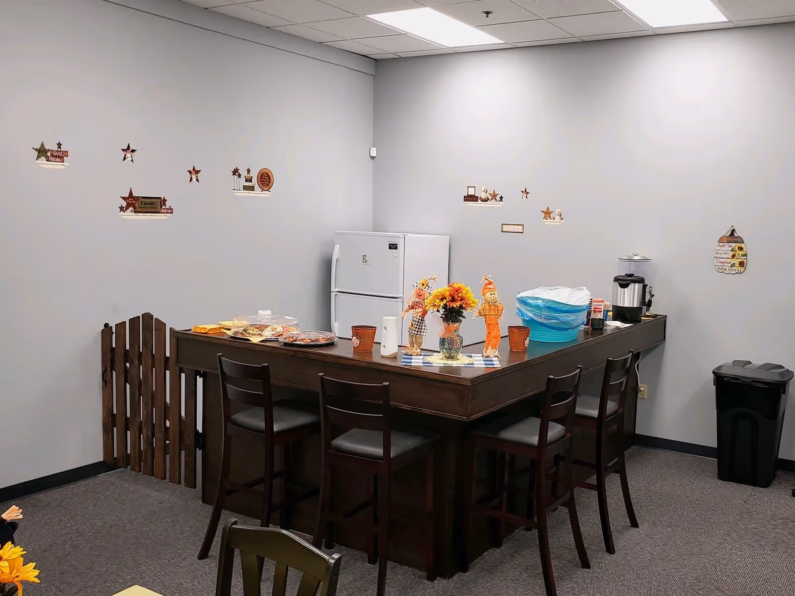 A small dining area with a dark wooden high table surrounded by four matching chairs. On the table are decorative items including a vase with sunflowers, two scarecrow figurines, covered food trays, paper towels, and a coffee urn. Behind the table is a white refrigerator and a small wooden gate attached to the wall. The walls are light gray and decorated with small autumn-themed wall hangings and stars. A black trash bin is placed in the corner of the room.