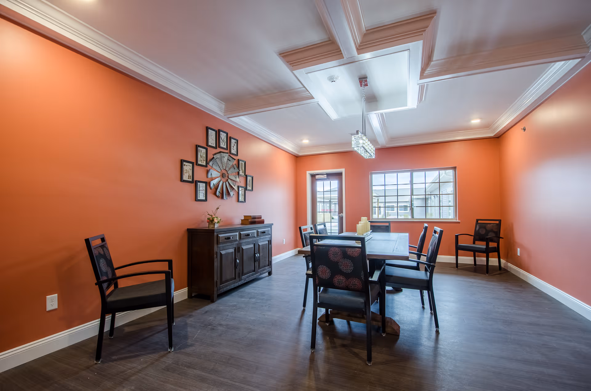 Bright dining room interior with orange walls, a rectangular table surrounded by chairs, a sideboard, and decorative wall art under a coffered ceiling.