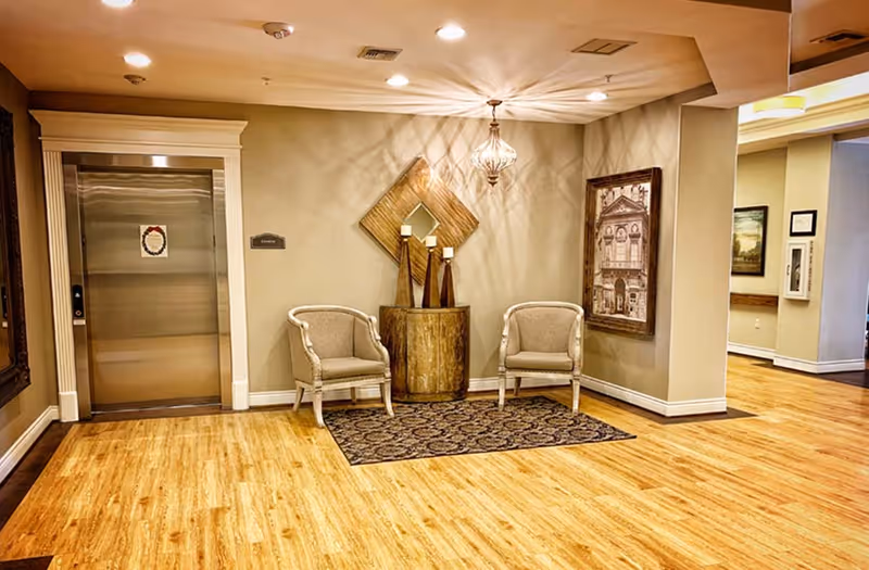 Interior view of a hallway area in an assisted living facility featuring a closed stainless steel elevator door framed by white trim on the left. Two beige upholstered chairs are placed on a patterned rug in front of a wooden side table with three candle holders. A decorative wall mirror and framed artwork hang on the beige walls. The floor is light wood, and ceiling lights illuminate the space warmly.