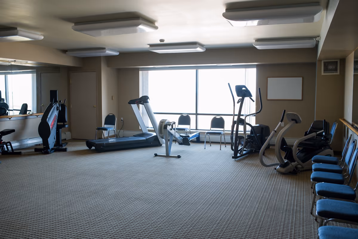 A fitness room with exercise equipment including a treadmill, rowing machine, elliptical trainer, and stationary bike. Several blue chairs are lined up along the walls. The room has a large window letting in natural light and a wall mirror on one side.
