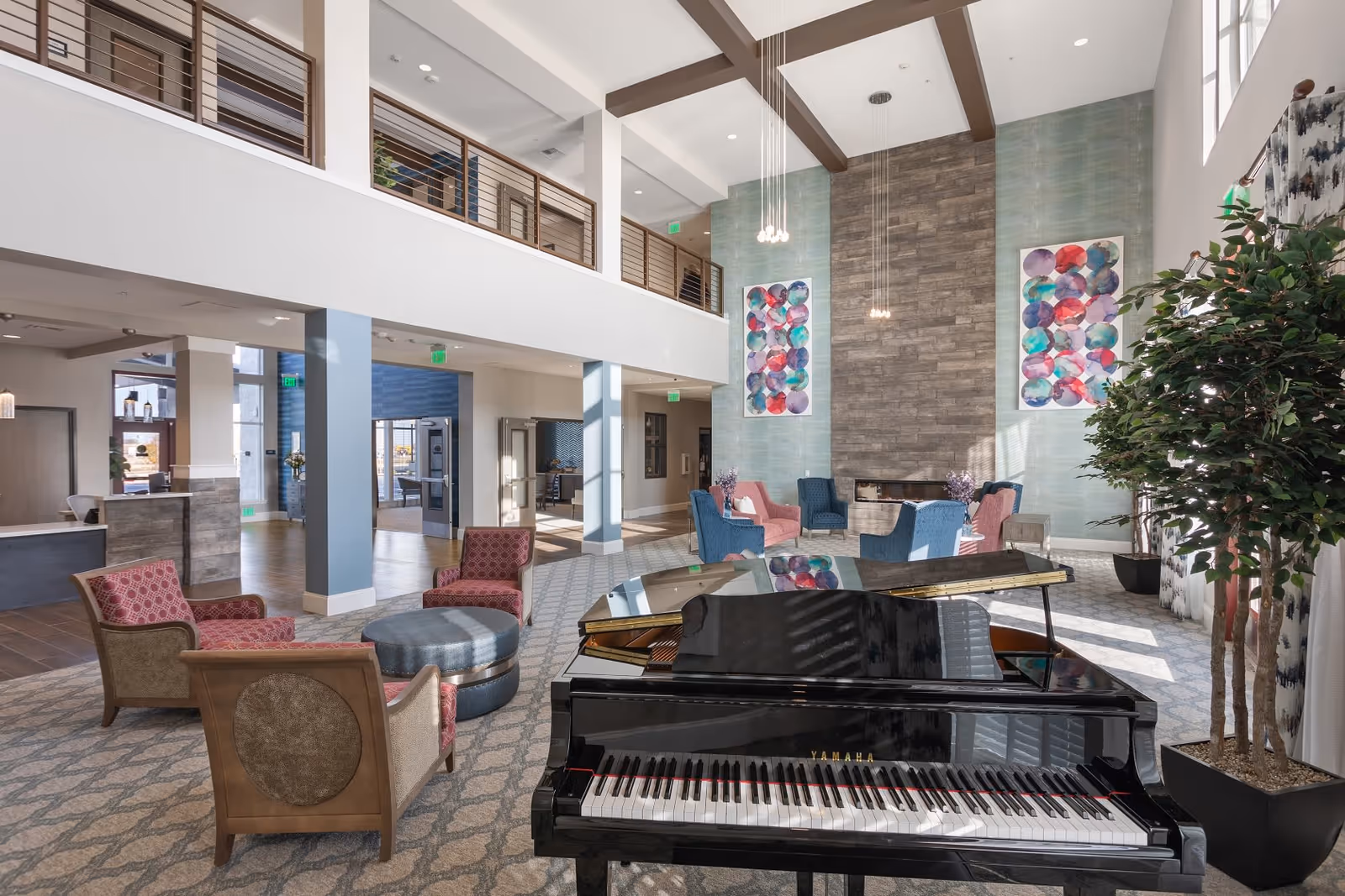 Spacious and well-lit senior living facility common area with a black Yamaha grand piano in the foreground, surrounded by comfortable seating including red and blue armchairs, a round ottoman, and a fireplace with modern art on the wall above it. The room features high ceilings with exposed beams, large windows allowing natural light, and a second-floor balcony overlooking the space.