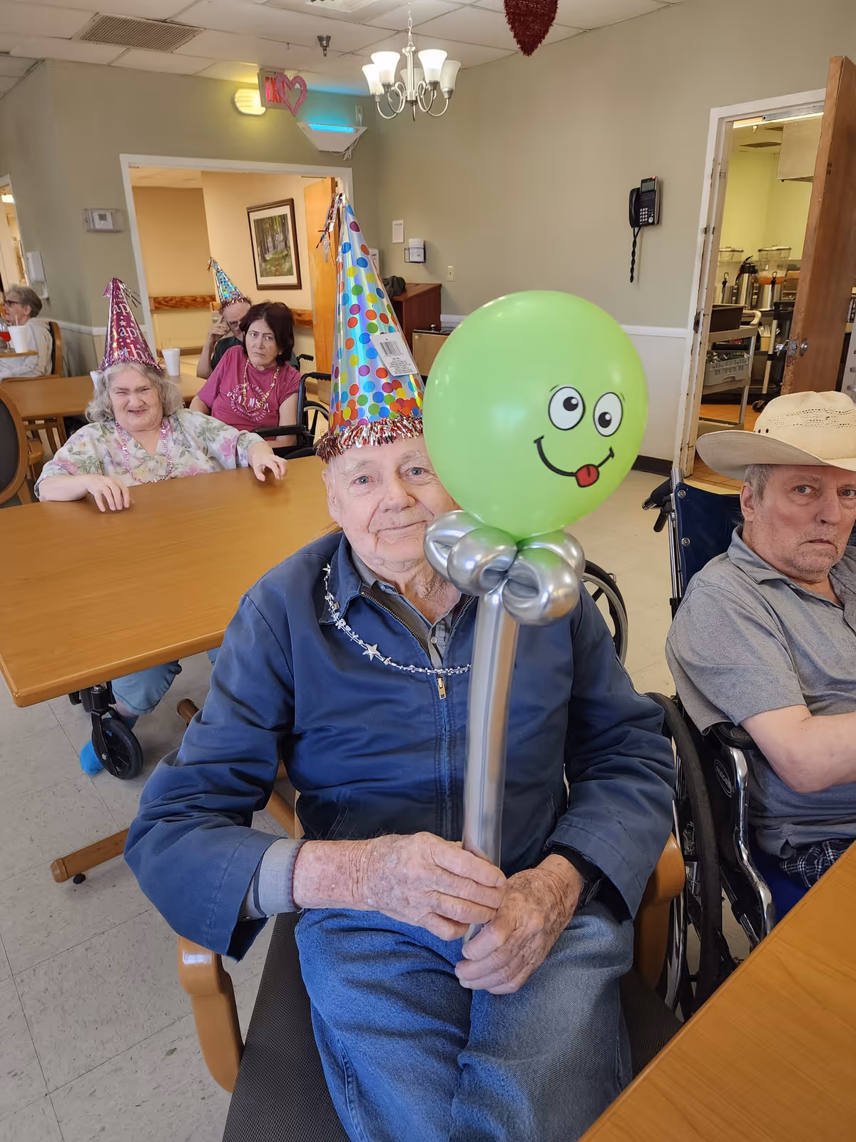 Elderly residents sitting at tables in a common room wearing colorful party hats. One man in the foreground holds a balloon sculpture with a green balloon featuring a smiling face. The room has light-colored walls, a chandelier, and an open door leading to a kitchen area.