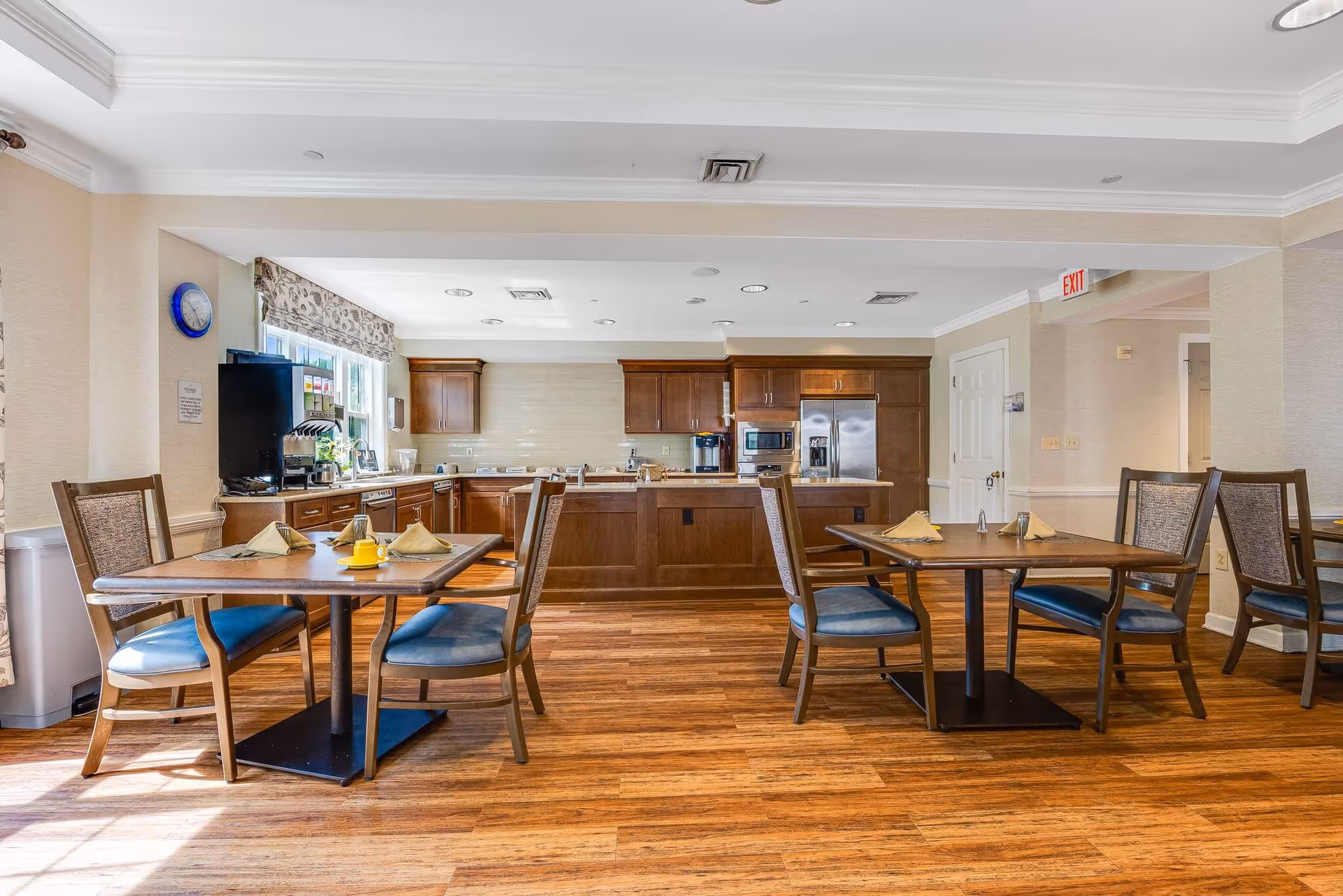 A bright dining area with wooden tables and chairs set with napkins and a yellow cup. The room features a kitchen in the background with wooden cabinets, a stainless steel refrigerator, microwave, and coffee machine. The floor is wooden, and there is a window with floral curtains letting in natural light.