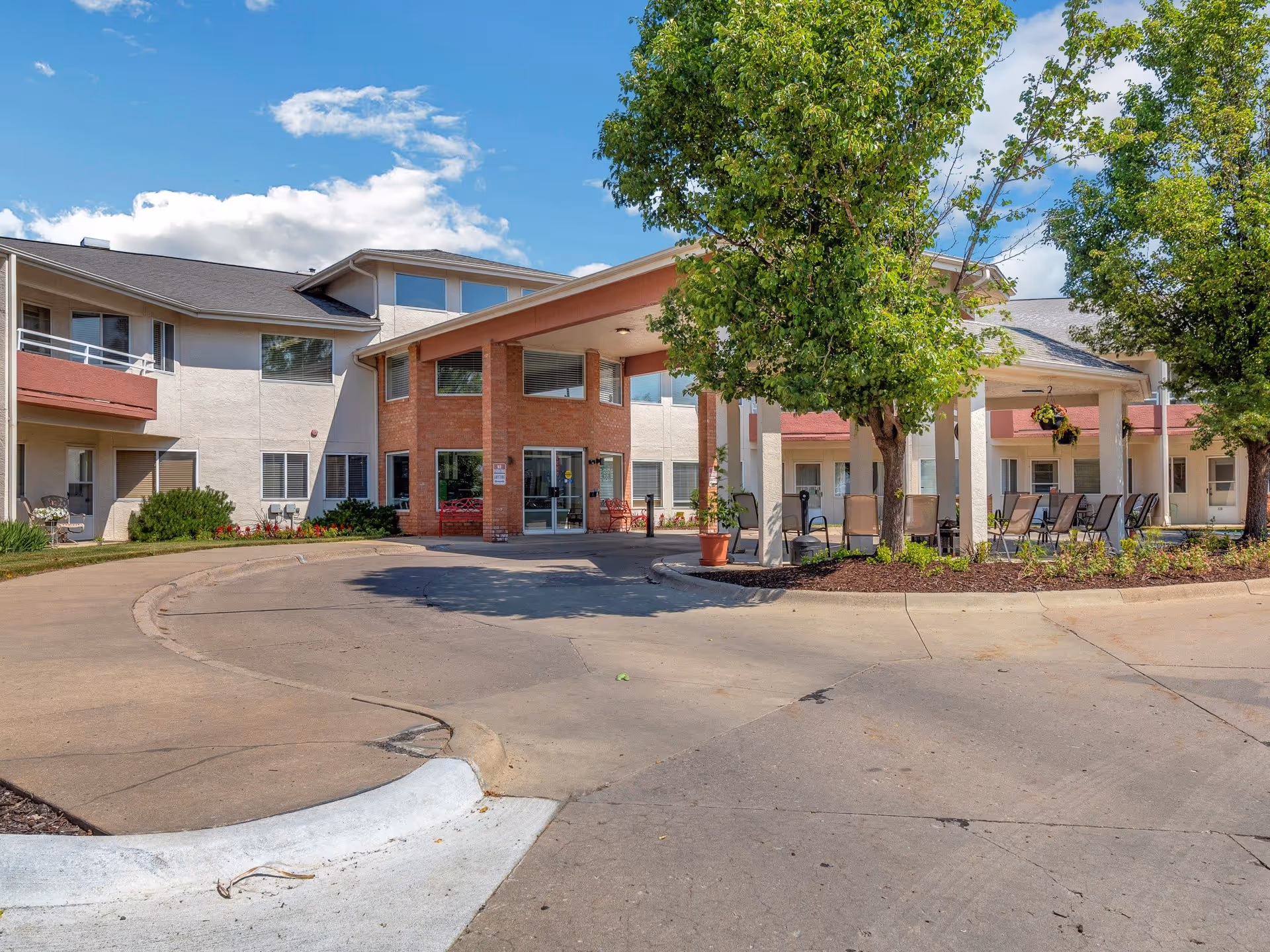 Exterior view of Walden Place Senior Living facility showing the main entrance with a covered drop-off area, surrounded by trees and outdoor seating. The building has a combination of brick and light-colored walls with multiple windows and balconies.