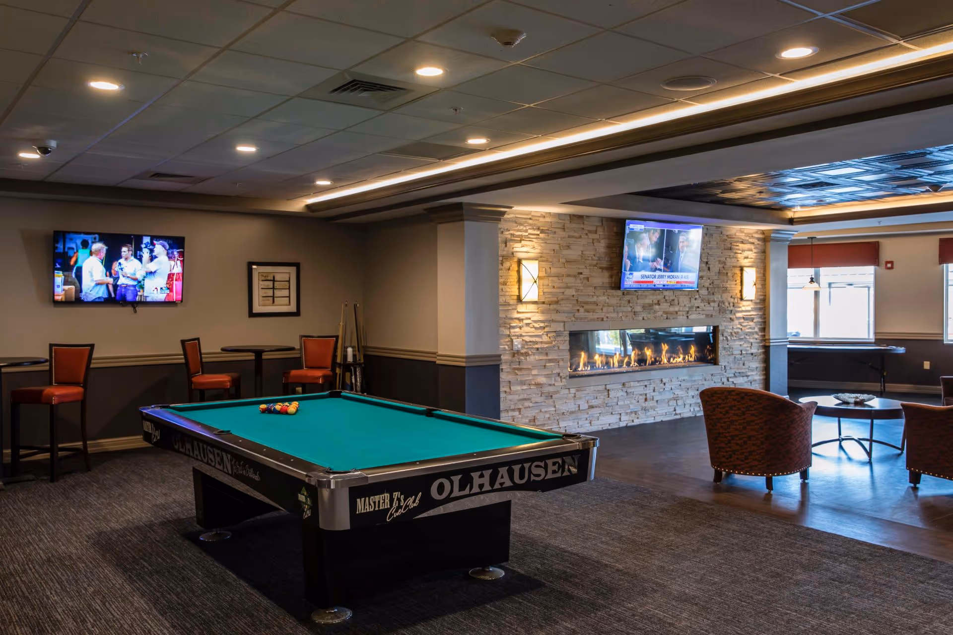 A recreational room featuring a pool table with billiard balls arranged on it, two wall-mounted televisions, a modern stone fireplace, several chairs and small tables, and a carpeted floor area.