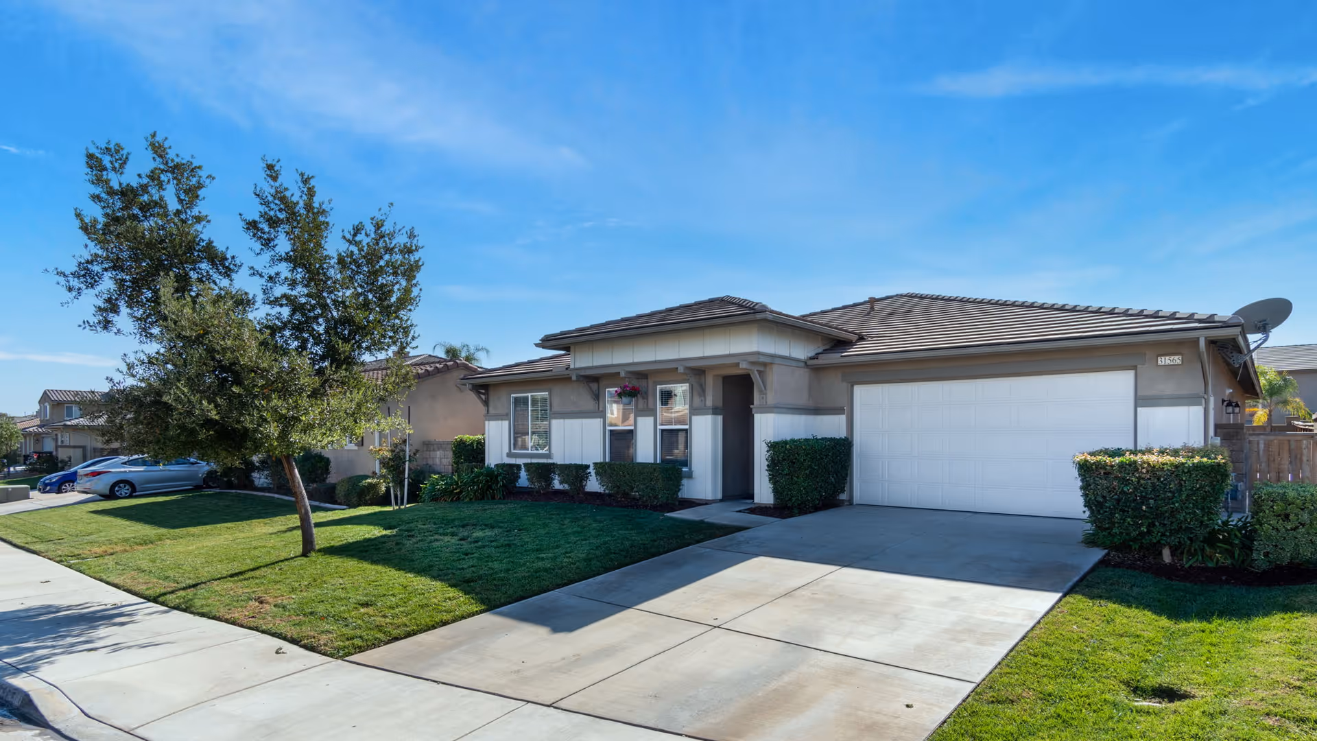 Single-story residential building with a two-car garage, a driveway, green lawn, and a tree in front under a clear blue sky.