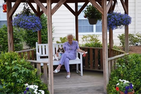 An elderly woman dressed in light purple sitting on a white bench inside a wooden gazebo, accompanied by a small dog. The gazebo is surrounded by green plants and hanging baskets with purple flowers. A building with white siding and windows is visible in the background.