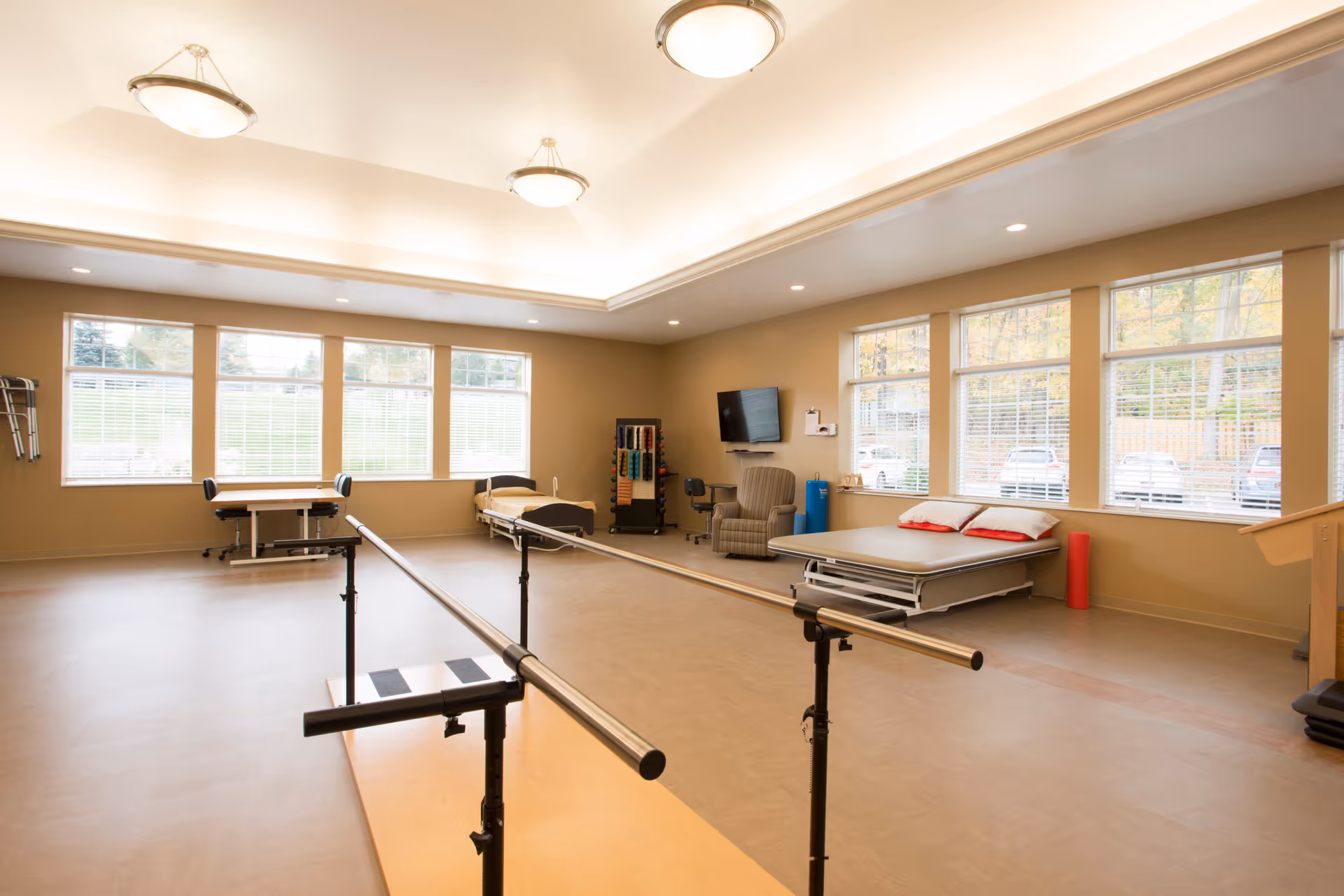 A spacious physical therapy room with large windows letting in natural light. The room contains parallel bars for walking exercises, a therapy bed with pillows, a recliner chair, a small desk with a chair, and various therapy equipment including weights and foam rollers. The walls are beige and the ceiling has recessed lighting and hanging light fixtures.