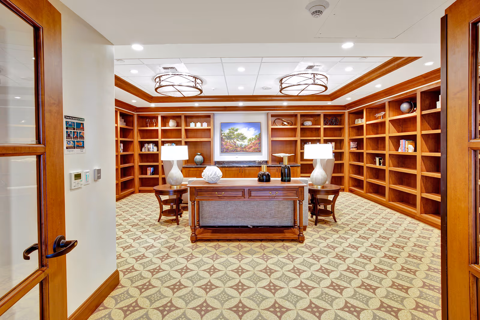 A spacious room with wooden bookshelves lining the walls, a patterned carpet, and a central wooden table with two lamps and decorative items. The ceiling has recessed lighting and two large circular light fixtures. A framed landscape painting hangs on the back wall.