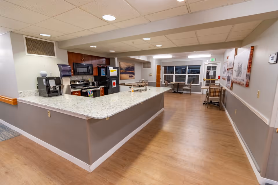 Interior view of a senior living facility kitchen and dining area with a large marble countertop island, stainless steel appliances including a stove and microwave, a coffee machine, and several tables and chairs near windows in the background.