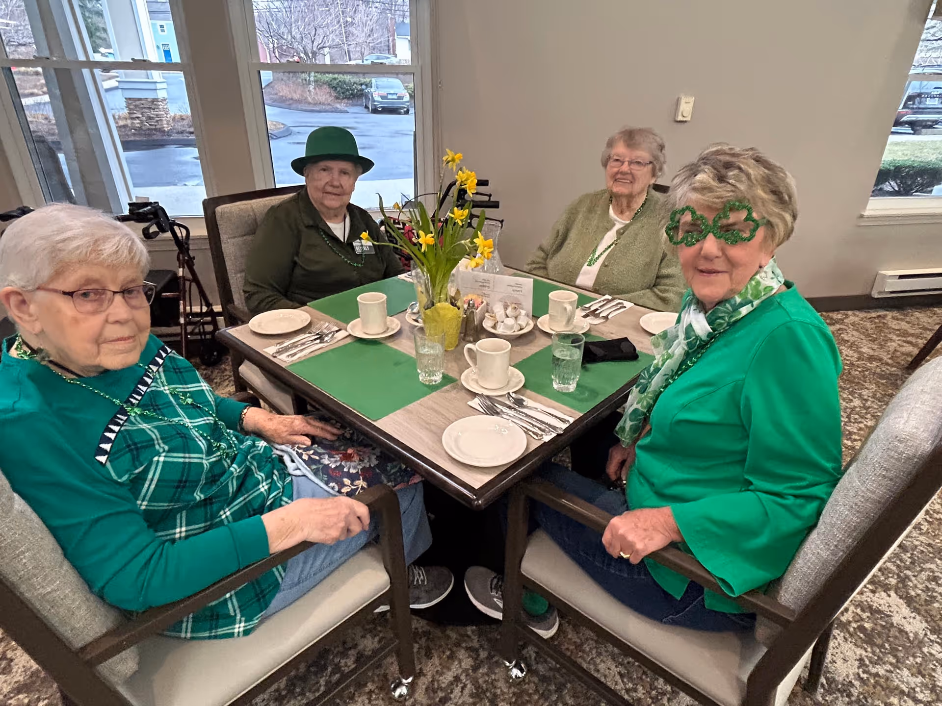 Four elderly women sitting around a square dining table set with plates, cups, and silverware. They are dressed in green clothing and accessories, with one woman wearing shamrock-shaped glasses and another wearing a green hat. A vase with yellow flowers is in the center of the table. The room has large windows showing an outdoor parking area.