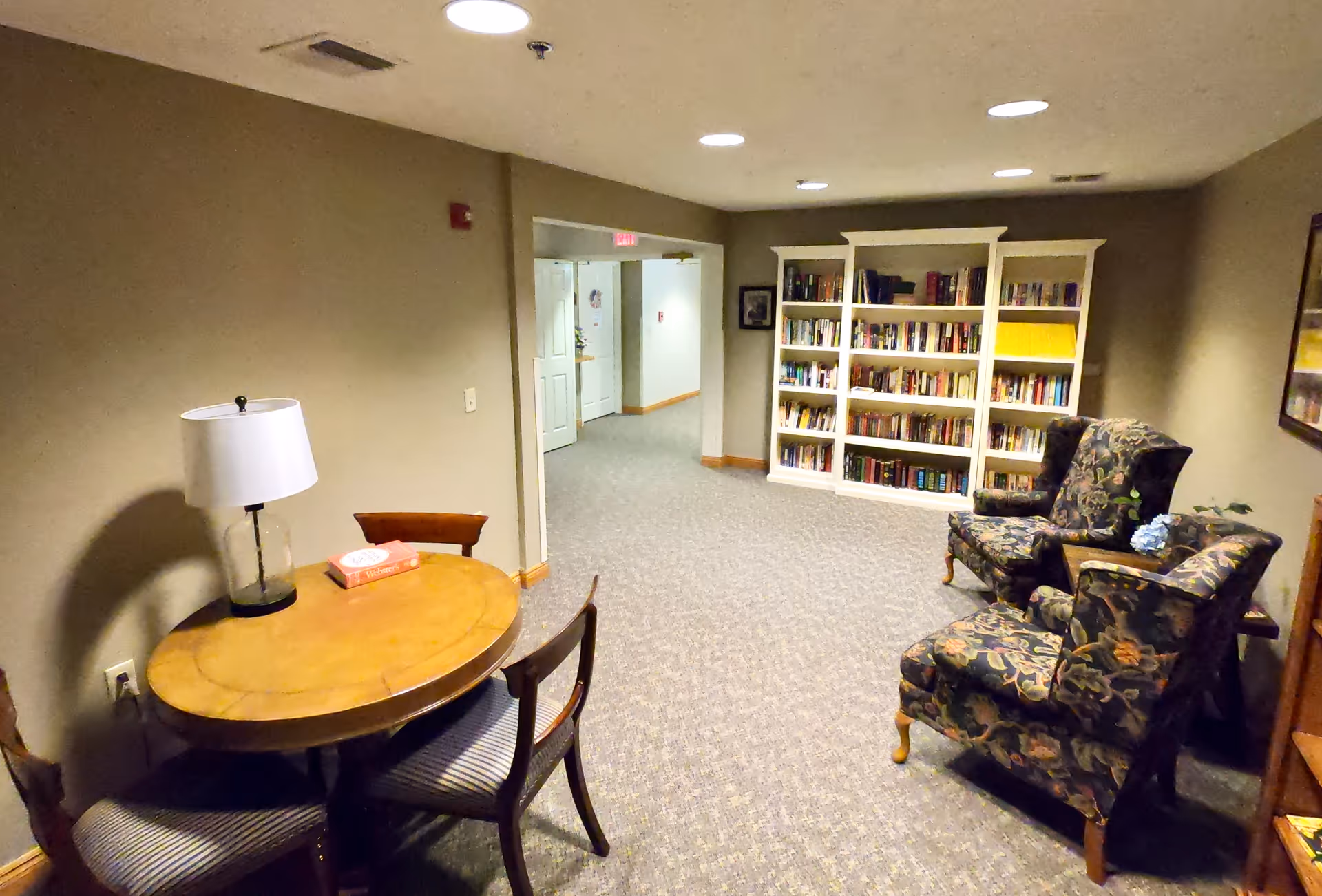 A cozy common area in an assisted living facility featuring a round wooden table with three chairs and a lamp, two floral upholstered armchairs, and a white bookshelf filled with books. The space has beige walls, carpeted flooring, and recessed ceiling lights.