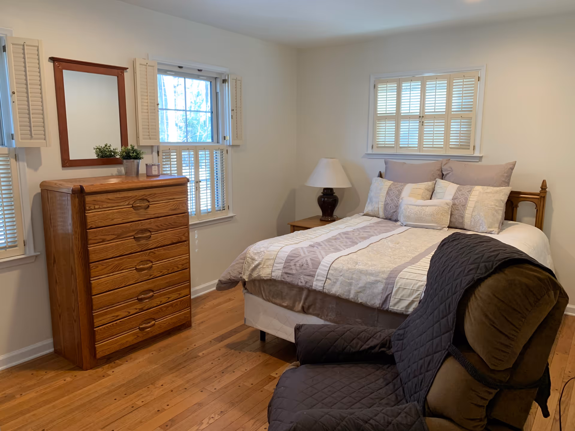 A cozy bedroom with a wooden dresser topped with a small plant and candle, a mirror above it, two windows with white shutters, a bed with multiple pillows and a patterned bedspread, and a cushioned armchair with a dark quilted cover.