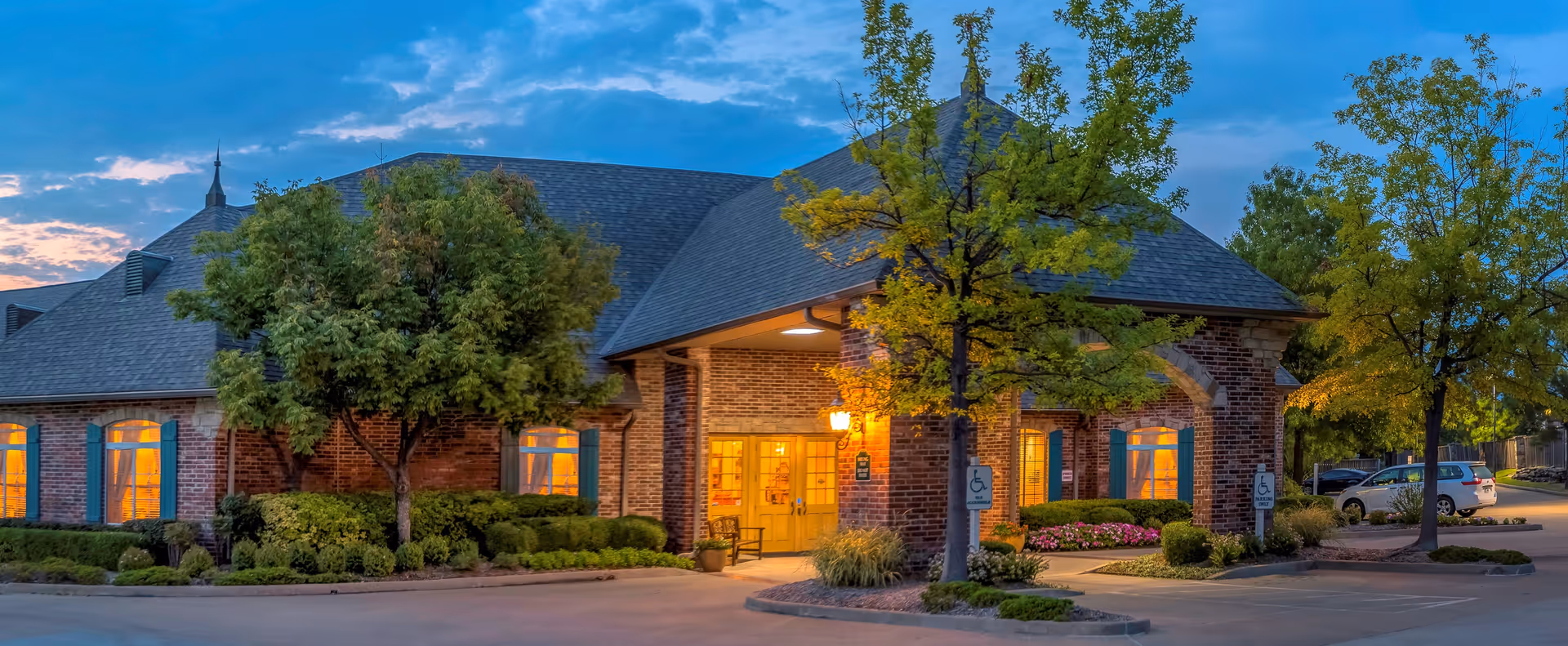 Exterior view of The Parke Assisted Living facility at dusk, showing a brick building with blue shutters, warm interior lights visible through the windows, trees and shrubs around the entrance, and a parking area with handicap parking signs.