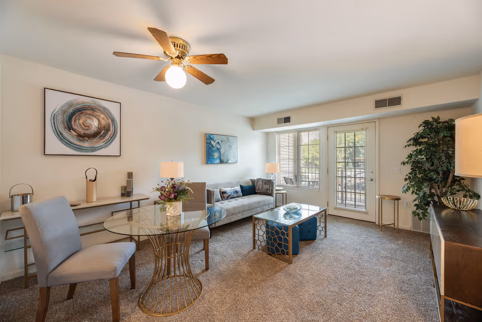 A bright and cozy living room with beige walls and carpet. The room features a ceiling fan with a light, a glass-top round table with two gray upholstered chairs, a beige sofa with decorative pillows, a glass coffee table with a geometric metal base, two table lamps, framed abstract artwork on the walls, a large potted plant, and a door with windows leading outside.