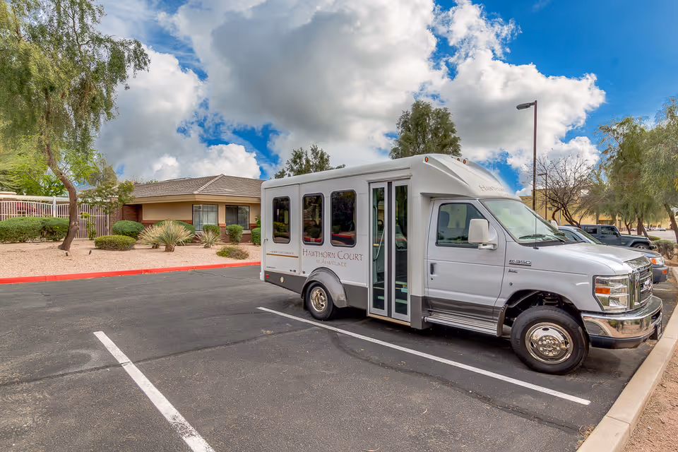 A Hawthorn Court at Ahwatukee Memory Care shuttle van parked in the facility parking lot in front of a low building and landscaped grounds under a cloudy sky.