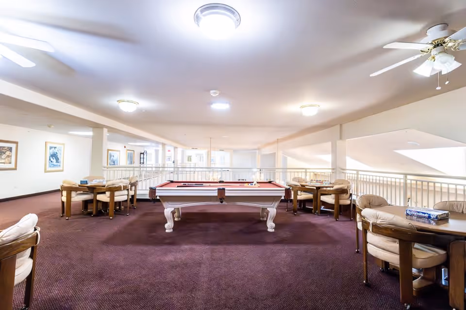A spacious recreational room with a red felt pool table in the center, surrounded by several round tables with cushioned chairs. The room has a carpeted floor, ceiling fans, and framed artwork on the walls. Large windows and railings overlook a lower level, allowing natural light to enter.