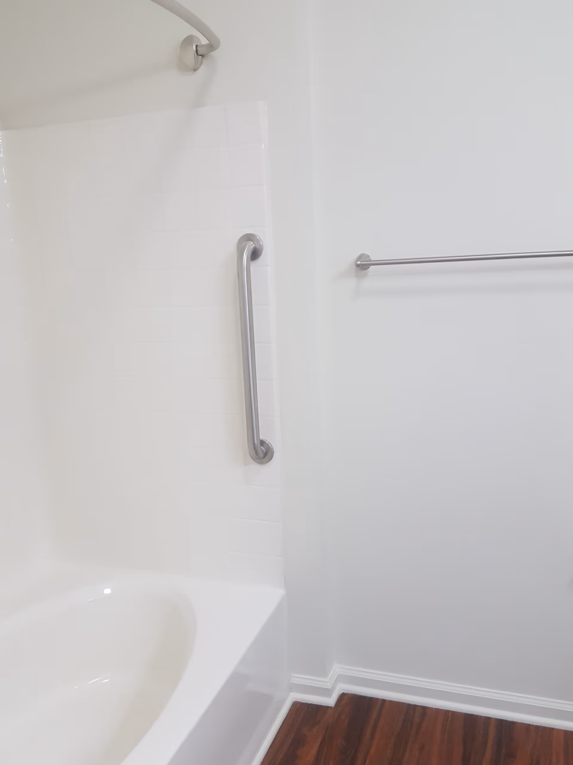 A clean bathroom corner featuring a white bathtub with a tiled wall surround. There is a silver grab bar mounted vertically on the tiled wall and a silver towel bar mounted on the adjacent white painted wall. The floor has dark wood-like flooring.