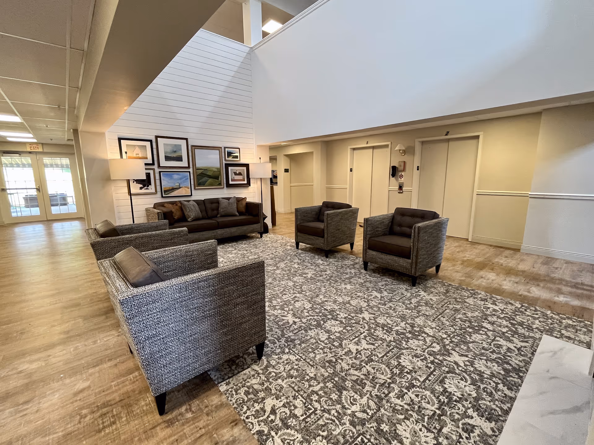 A spacious and well-lit lounge area with a patterned rug, two armchairs, a sofa, and two floor lamps. The walls are decorated with framed artwork, and there are two elevators visible in the background. The floor is wooden, and there is a high ceiling with a partial second floor visible.