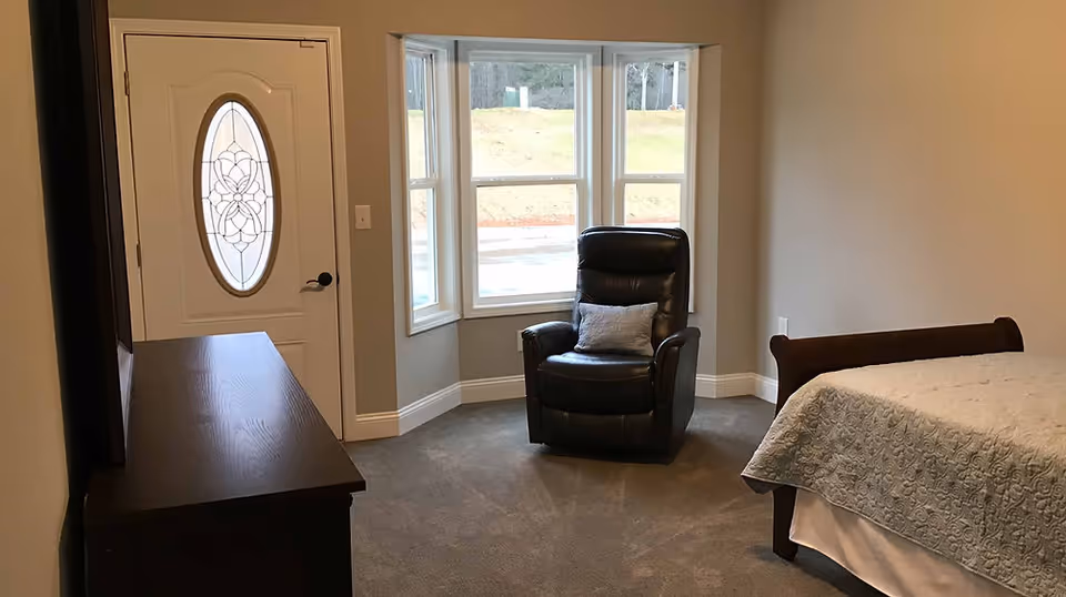 A bedroom with a large window featuring three panels, a black leather recliner with a gray pillow, a wooden dresser, a bed with a gray quilt, and a white door with an oval decorative glass panel.