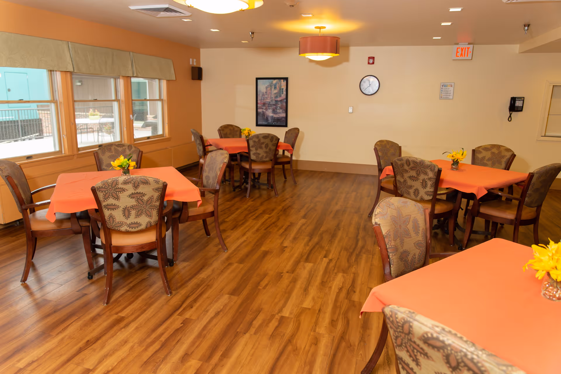 A dining room with several tables covered in orange tablecloths, each surrounded by patterned chairs. Small yellow flower arrangements are placed on each table. The room has wooden flooring, large windows on one side, a wall clock, a framed picture, and an exit sign above a door.