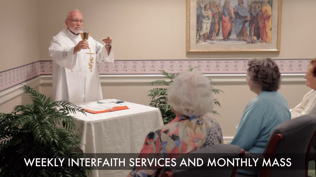 A priest dressed in white robes is holding a chalice and a communion wafer in front of a small group of elderly women seated in chairs, participating in a religious service inside a room with a framed religious painting on the wall and potted plants nearby.