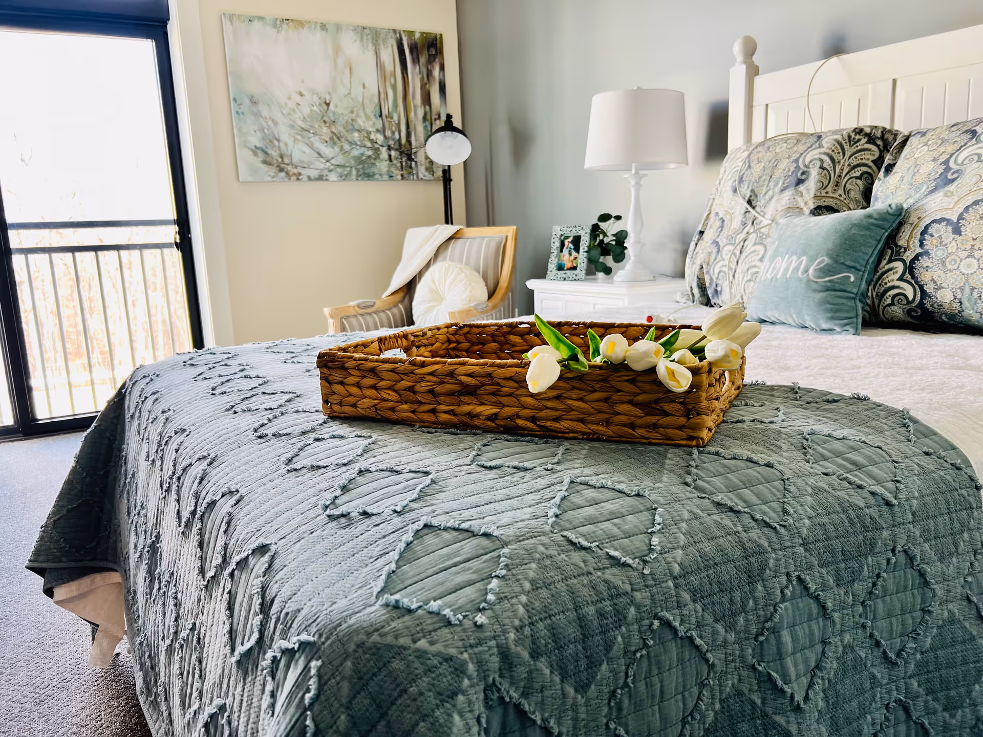A cozy bedroom with a neatly made bed covered in a textured blue-green quilt. On the bed is a woven basket holding white tulips. Behind the bed are decorative pillows, including one with the word 'Home'. To the right is a white nightstand with a lamp, a framed photo, and a small plant. In the background, there is a wooden chair with a cushion and a throw blanket, a floor lamp, and a large window or sliding glass door letting in natural light.
