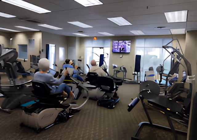 A fitness room with several exercise machines including stationary bikes and weight machines. Three elderly individuals are seated on the exercise bikes, facing a wall-mounted television. The room has large windows with blinds and overhead fluorescent lighting.