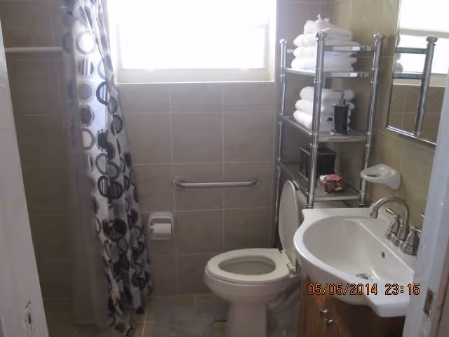 Small bathroom with beige tiled walls and floor, featuring a white toilet, a white sink with a silver faucet, a metal shelving unit stocked with folded white towels and toiletries, a shower area with a curtain decorated with black and white circular patterns, and a window letting in natural light.