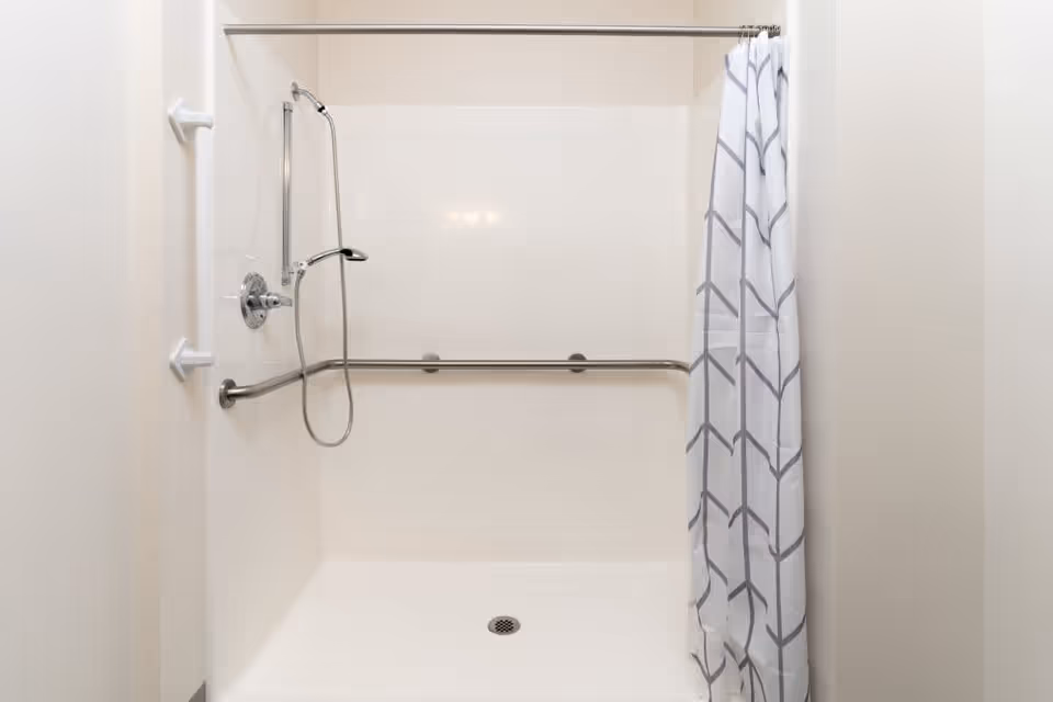 A clean, accessible shower stall with white walls, a handheld showerhead, and two horizontal grab bars. A white shower curtain with a gray geometric pattern is partially drawn to the right.