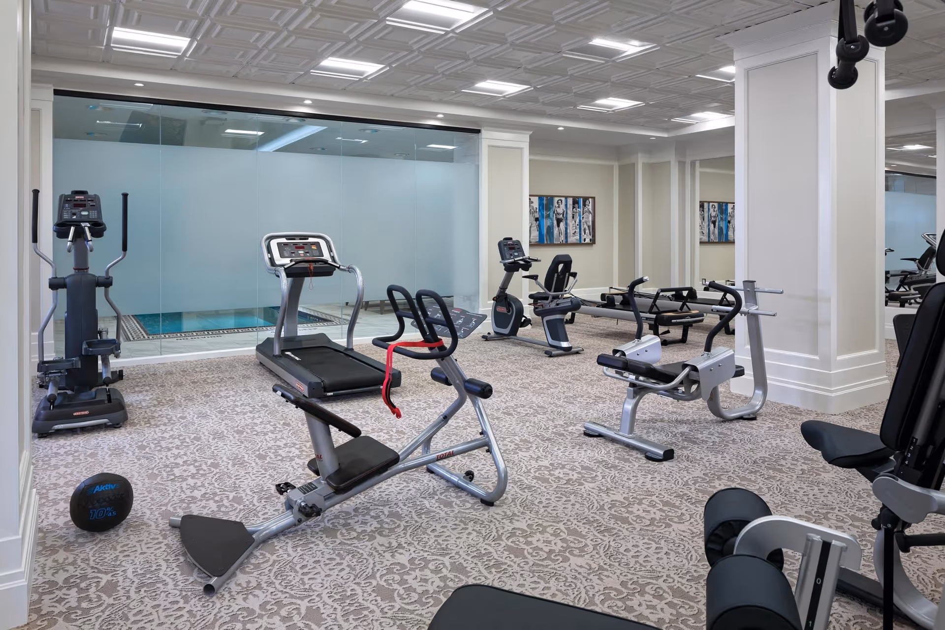 A well-lit fitness room with various exercise equipment including a treadmill, elliptical machine, stationary bike, and weight machines. The room has patterned carpet flooring, white paneled walls, and a large glass window revealing an indoor pool area. There are framed artworks on the walls and a medicine ball on the floor.