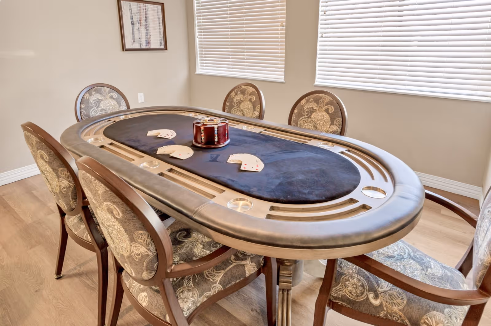 A poker table with a black felt surface and built-in cup holders surrounded by six upholstered chairs with patterned fabric. The room has light-colored walls, two windows with blinds, and a framed picture on the wall.