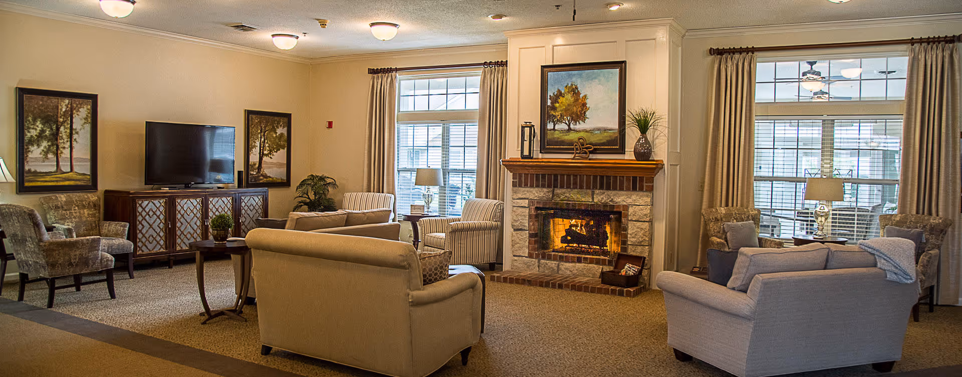 A cozy living room area in a senior living facility featuring a lit fireplace with a painting above it, several upholstered armchairs and sofas arranged around the room, a flat-screen TV on a wooden cabinet, framed artwork on the walls, large windows with curtains, and table lamps providing warm lighting.