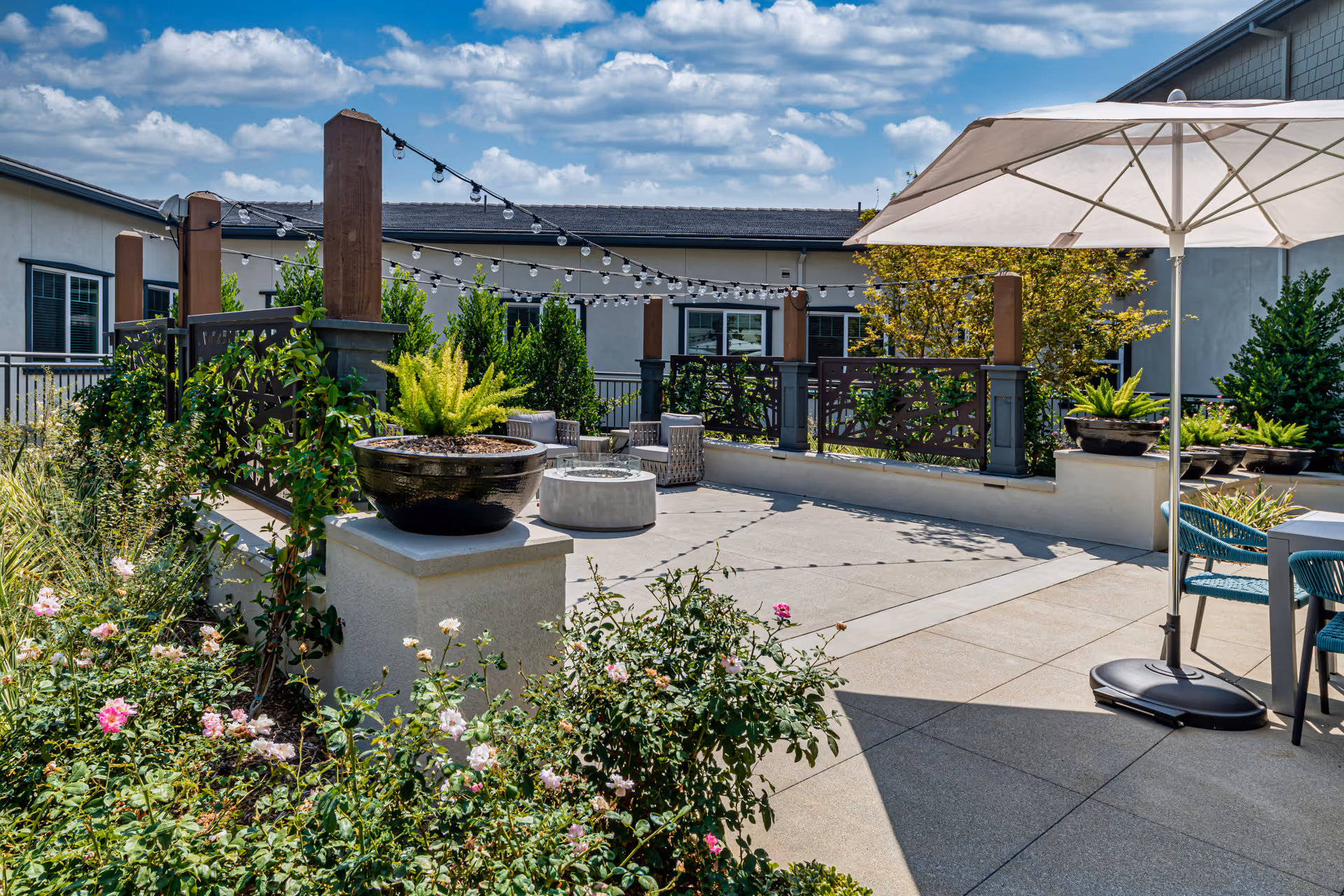 Outdoor patio area at Allara Senior Living featuring a concrete floor, potted plants, string lights hanging between wooden posts, a large white patio umbrella, and outdoor seating including chairs and a table. The sky is partly cloudy and the surrounding building walls are visible.
