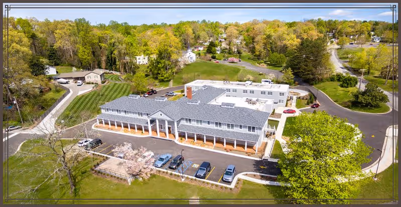 Aerial view of Alexander Guest House, a large two-story building with a gray roof and white columns along the front porch. The building is surrounded by green lawns, trees, and a parking lot with several cars. The area around the facility is lush with trees and residential houses in the background.