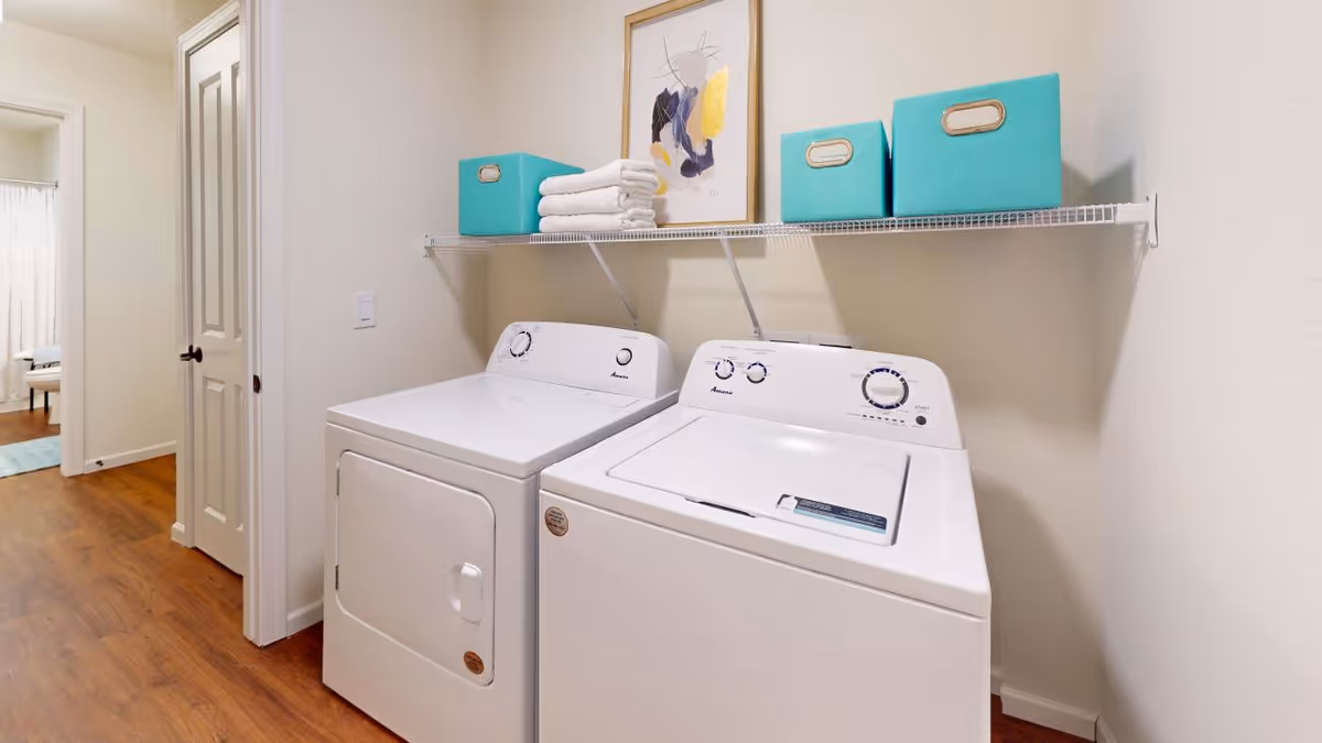A laundry area with a white washer and dryer beneath a wire shelf holding folded towels, teal storage boxes, and wall art.