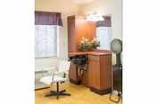 Interior salon area with a styling chair, wash basin, mirror, and wooden cabinetry beside a window.