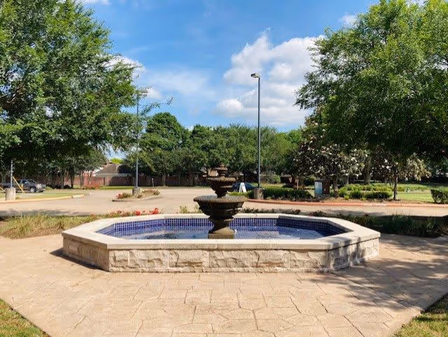 Outdoor scene featuring a stone fountain with a blue tiled interior, surrounded by a paved area. There are green trees and shrubs around the fountain, with a clear blue sky and some clouds above.