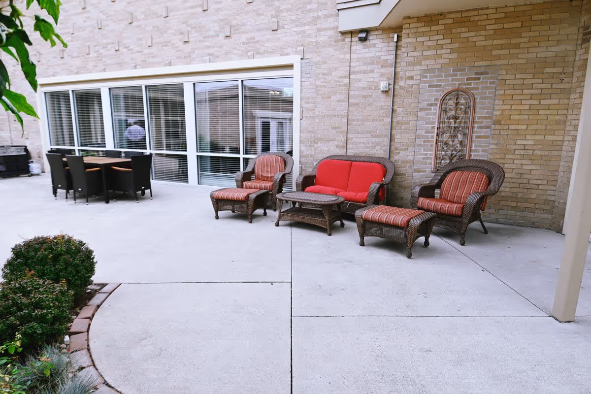 Outdoor patio area with wicker furniture including two armchairs with red and striped cushions, a loveseat with red cushions, a small coffee table, and a dining table with six black chairs. The patio is adjacent to a brick building with large windows and some greenery on the left side.