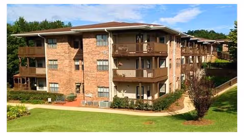 Exterior view of a three-story brick residential building with balconies, surrounded by green grass, trees, and a clear blue sky.